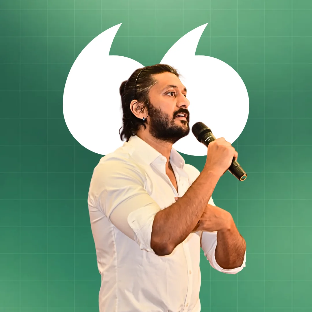 Chetan Ahimsa speaking into a microphone, in a white shirt, against a forest-green background framed by large white quotation marks
